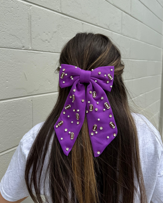 Person with long hair wearing a purple hair bow with gold accents against a light gray wall.