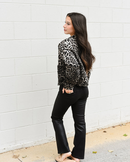 Woman wearing a leopard print jacket and black pants against a white wall.