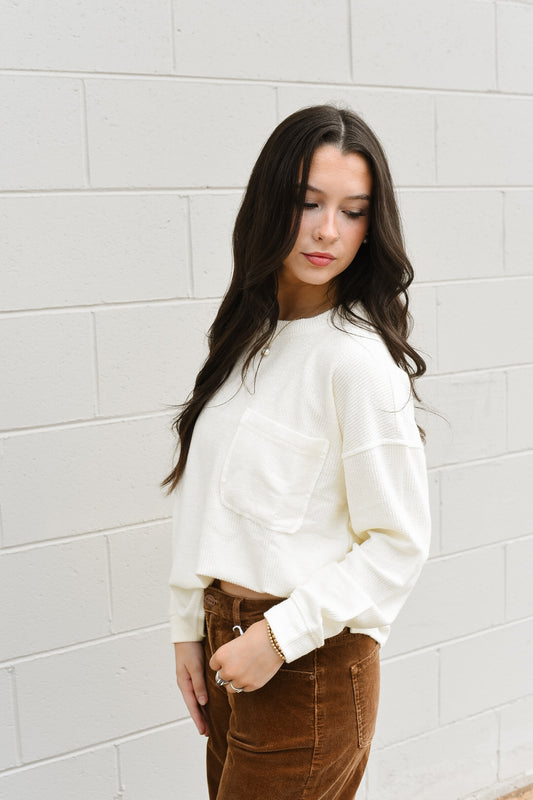 Woman wearing a white sweater and brown pants against a gray brick wall