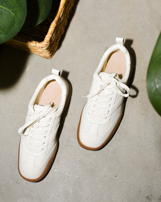 Pair of white sneakers with brown soles on a concrete floor.