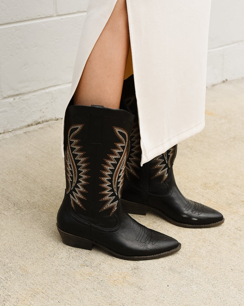 Black cowboy boots with intricate patterns worn by a person against a white tiled wall.