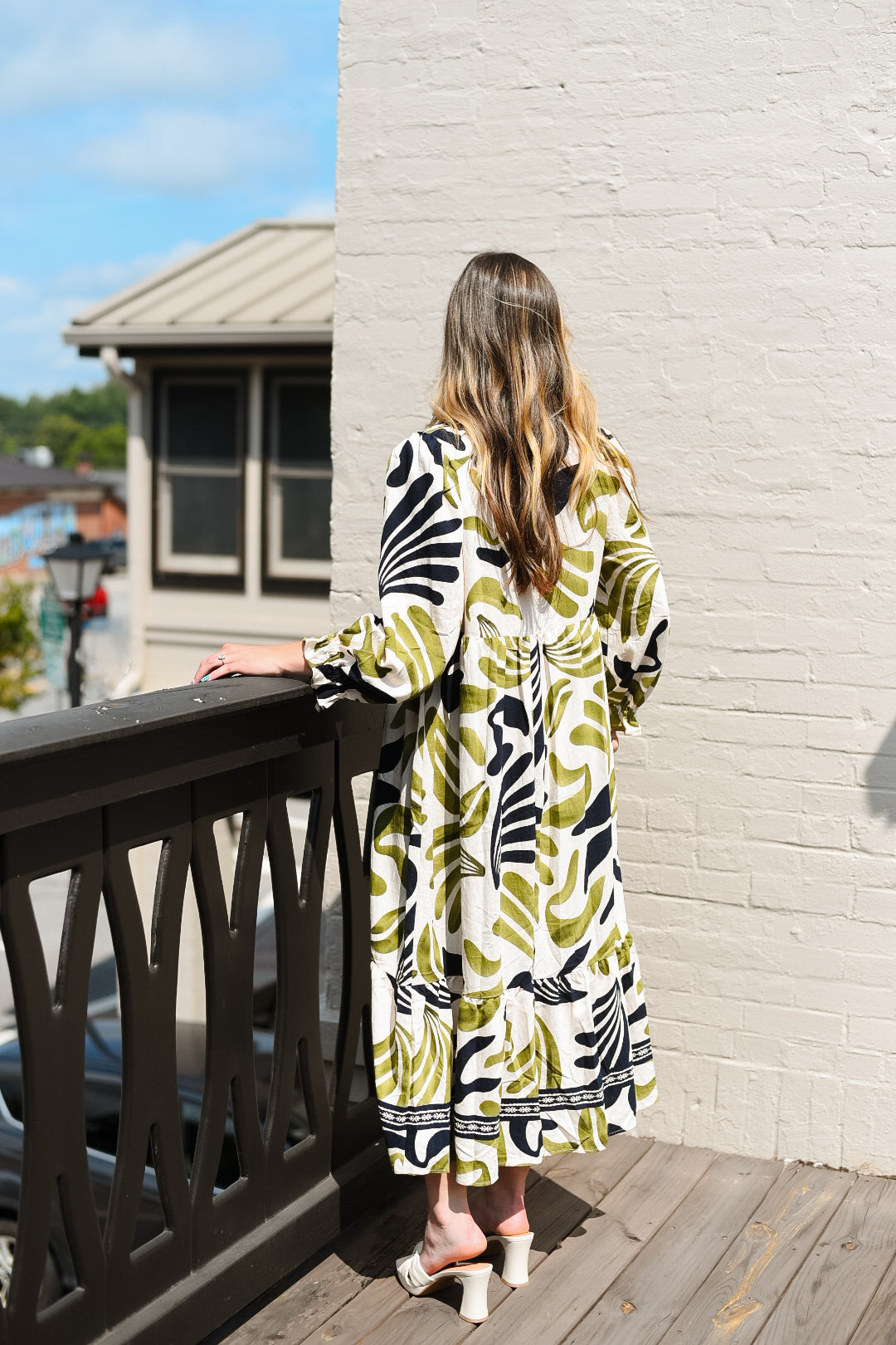 Woman in a patterned dress standing on a balcony with a brick wall and building in the background.