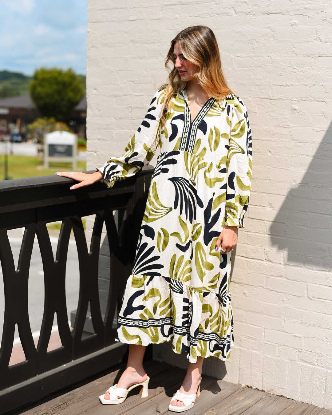 Woman in a patterned dress standing on a wooden deck with a white brick wall and greenery in the background.