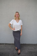 A woman standing against a gray background, wearing a white v-neck t-shirt with short sleeves and a black skirt.