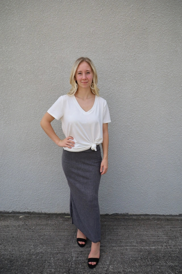 A woman standing against a gray background, wearing a white v-neck t-shirt with short sleeves and a black skirt.