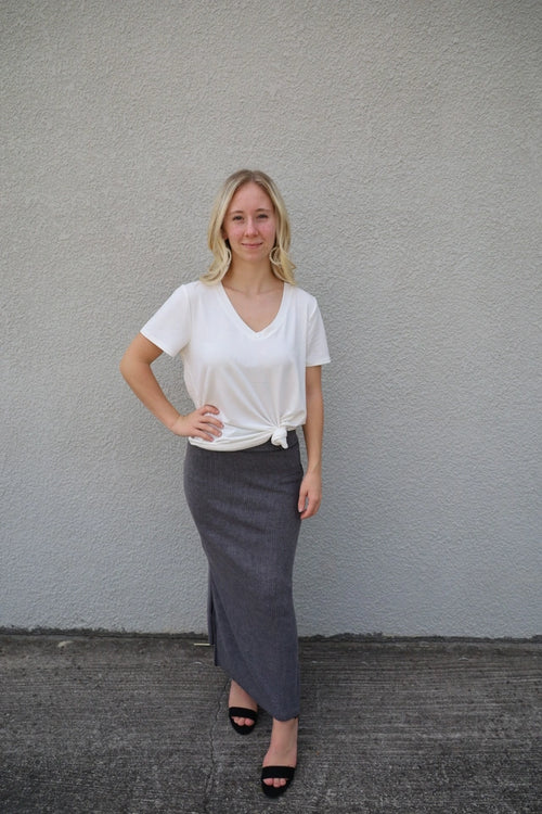 A woman standing against a gray background, wearing a white v-neck t-shirt with short sleeves and a black skirt.