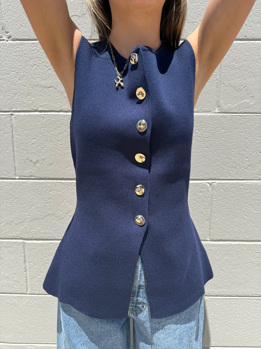Person wearing a navy blue sleeveless top with gold buttons against a light gray brick wall.