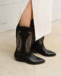 Black cowboy boots with intricate patterns worn by a person against a white tiled wall.