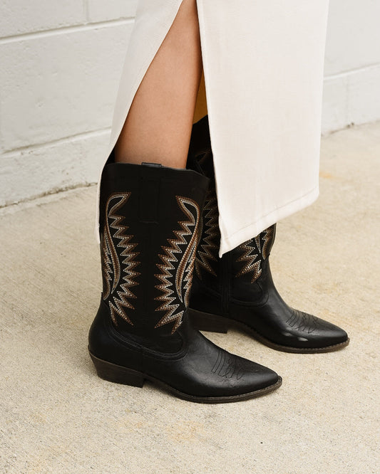 Black cowboy boots with intricate patterns worn by a person against a white tiled wall.