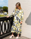 Woman in a patterned dress standing on a wooden deck with a white brick wall and greenery in the background.