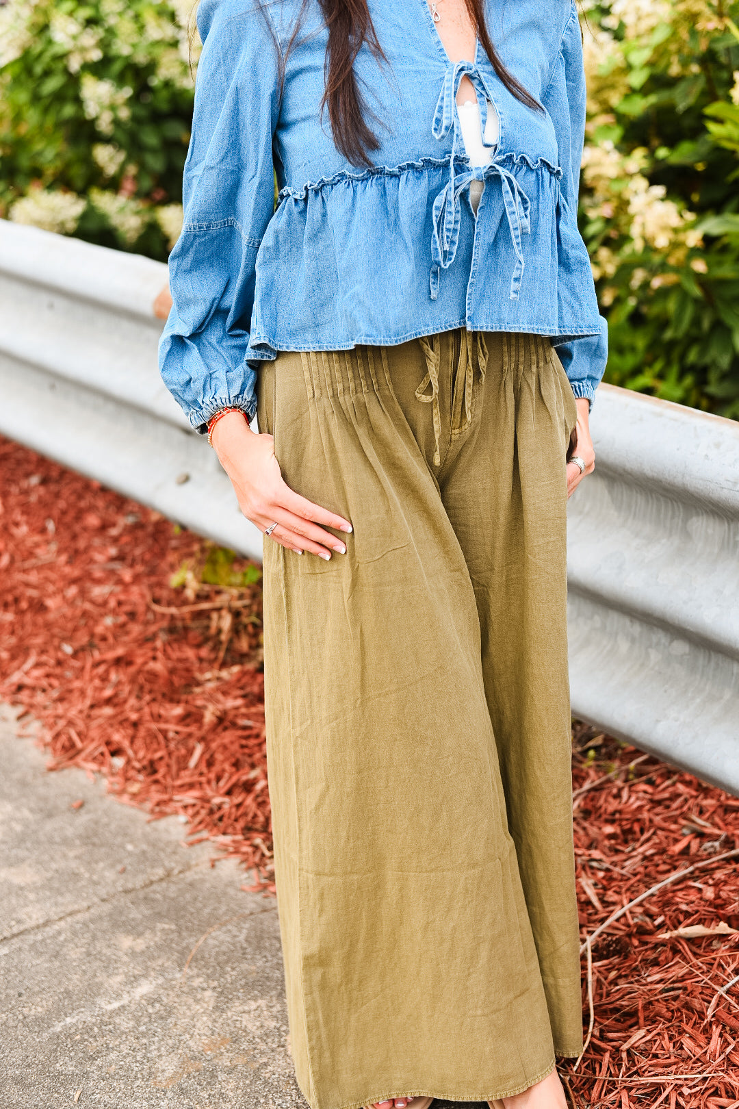 Person wearing a blue top and green pants standing outdoors with plants in the background
