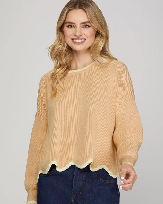 Woman wearing a beige sweater with scalloped hem and dark blue jeans on a gray background